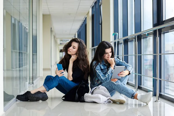 两个年轻的黑发女孩 坐在机场走廊的地板上 背着行李 检查电话和平板电脑 穿着休闲装 女朋友 坐飞机 等飞机 — 图库照片