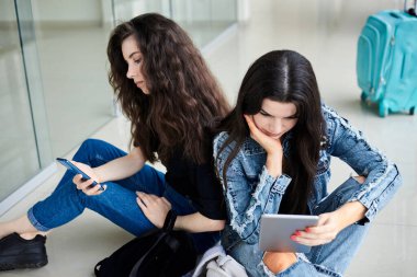 İki genç esmer kız, açık hava koridorunda arkada bavullarıyla oturuyorlar, telefon ve tableti kontrol ediyorlar, günlük kot giyiyorlar. Kız arkadaşlar, hava yoluyla seyahat ediyorlar, uçmayı bekliyorlar..