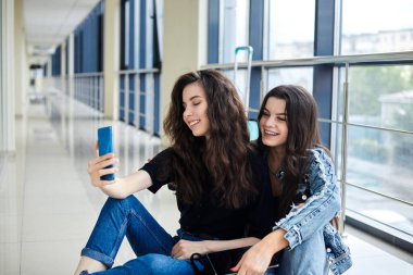İki genç esmer kız, açık hava koridorunda arkada bavullarıyla oturuyorlar, telefonla selfie çekiyorlar, gülümsüyorlar, günlük giysiler giyiyorlar. Kız arkadaşlar, hava yoluyla seyahat ediyorlar, uçmayı bekliyorlar..