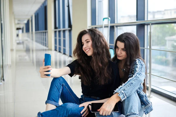 两个年轻的黑发女孩 坐在机场走廊的地板上 背着行李 拿着手机自拍 面带微笑 穿着休闲装 女朋友 坐飞机 等飞机 — 图库照片