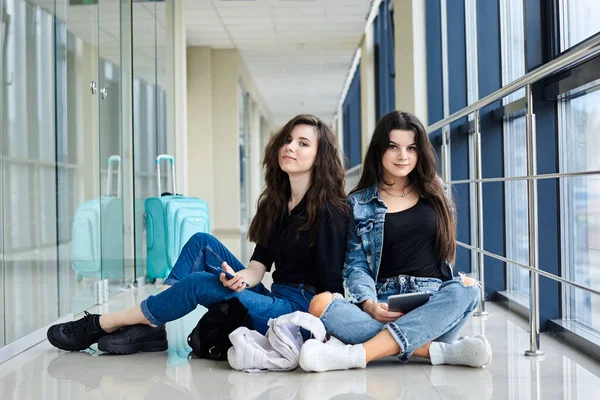 2人の若いブルネットの女の子は 明るい空港の廊下の床に 荷物を背後に タブレットを保持し 笑顔で カジュアルなジーンズの服を着て座っている 女の子の友達 飛行機で旅行 フライトを待っている — ストック写真