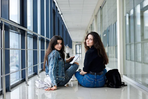 2人の若いブルネットの女の子は 光の空港の廊下の床に カメラに背を向けて カジュアルなジーンズの服を着て座っている 女の子の友達 飛行機で旅行 フライトを待っている — ストック写真