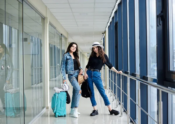 Two young brunette girls, standing in light airport hallway with small carry on luggage,,wearing casual jeans clothes, holding white and black backpacks.Girlfriends, traveling by air.