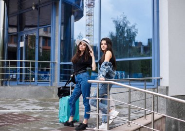 İki genç esmer kız, günlük kot giyiyorlar, mavi modern cam binanın önünde nane bavullarıyla duruyorlar kız arkadaşları geziyorlar, yürüyüşten sonra dinleniyorlar, otobüs durağında tren bekliyorlar..