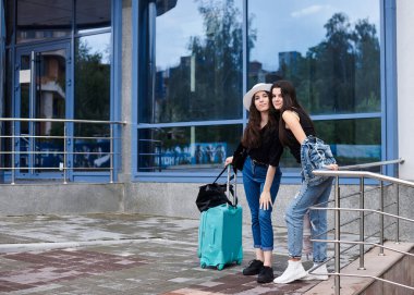 İki genç esmer kız, günlük kot giyiyorlar, mavi modern cam binanın önünde nane bavullarıyla duruyorlar kız arkadaşları geziyorlar, yürüyüşten sonra dinleniyorlar, otobüs durağında tren bekliyorlar..