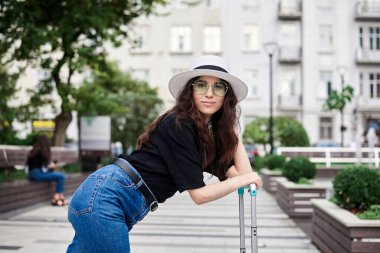 Genç esmer kadın, siyah gömlek, kot pantolon ve yeşil güneş gözlüklü beyaz şapka giyiyor, nane şekerine yaslanıyor, yazın eski tarih binasının önünde yürüyor. Şehirde gezgin.