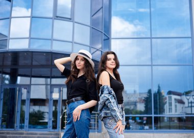 İki genç esmer kız, günlük kot giyiyorlar, mavi cam binanın önünde duruyorlar. Kız arkadaşları şehirde geziyor, sırt çantaları taşıyor, etrafı geziyor. Etkin yaşam tarzı kavramı.
