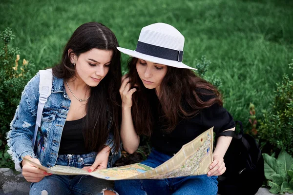 两个年轻的黑发女孩 穿着休闲装 坐在绿草上 拿着旅行地图 在城里查看路线 城市观光游览 旅游生活方式 — 图库照片