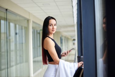 Genç esmer kadın, beyaz pantolon ve siyah bluz giyiyor, telefonu tutuyor, açık ofis binasının penceresinde dikiliyor. Ofis çalışanı tatilde..