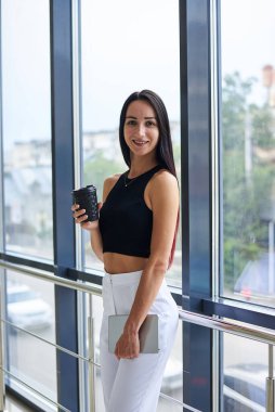 Genç esmer kadın, beyaz pantolon ve siyah bluz giyiyor, kahve fincanı tutuyor, açık ofis binasının camına yaslanıyor. İş kadını, şirket toplantısına hazırlanıyor. Ofis çalışanı tatilde..