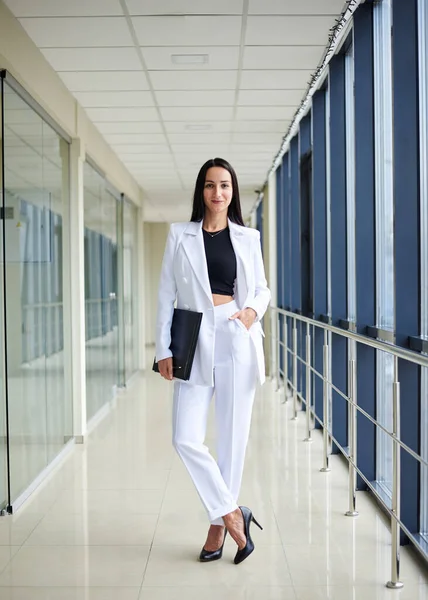 Young brunette woman, wearing white suit, holding black folder with documents,standing in white office building with huge windows.Businesswoman, getting ready for corporate meeting.Business relations