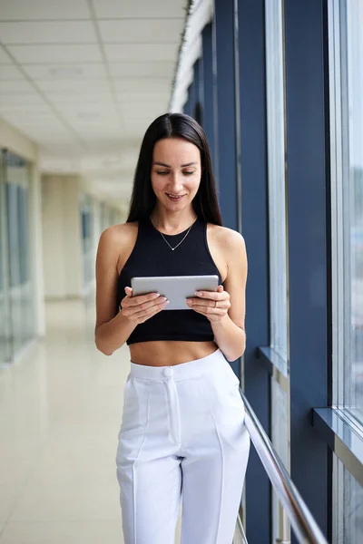 stock image Young brunette woman, wearing white pants and black top, holding silver tablet, standing in light office building. Businesswoman, getting ready for corporate meeting. Office worker on break.