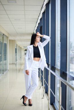 Genç esmer kadın, beyaz pantolon ve ceket giyiyor, açık koridorda duruyor, sosyal medya için poz veriyor, pencereye yaslanıyor. İş kadını molada. Kadın portresi fotoğrafı..