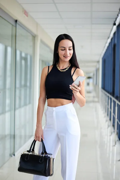 若いブルネットの女性は 白いパンツと黒い上を着て 携帯電話を見て 携帯電話のオフィスビルに立って黒い財布を持っている ビジネスマンは 企業の会議の準備をする — ストック写真