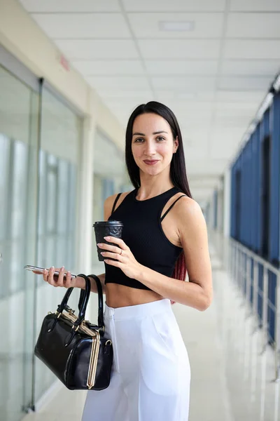 若いブルネットの女性は 白いパンツと黒の上を着て オフィスビルに立って 黒い財布 携帯電話 紙のコーヒーカップを保持する 昼休み中のビジネスマン — ストック写真