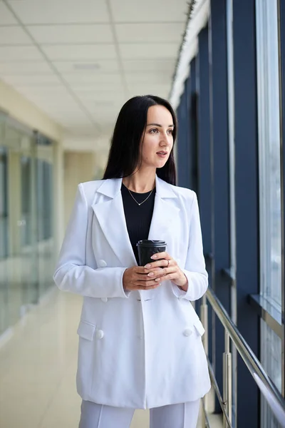 Young brunette woman, wearing white business suit, holding black paper coffee cup, standing in light office building near window. Businesswoman on lunch break. Corporate culture concept.