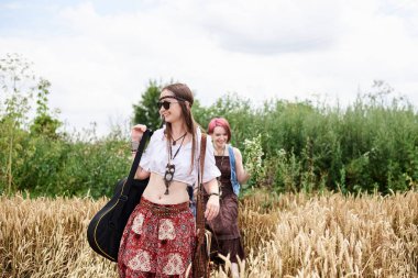 İki hippi kadın, boho tarzı kıyafetler giyiyorlar, gitar tutuyorlar, buğday tarlasına yürüyorlar, gülümsüyorlar. Kırsalda birlikte seyahat eden bayan arkadaşlar. Eko turizm konsepti. Yaz tatili boş zaman.