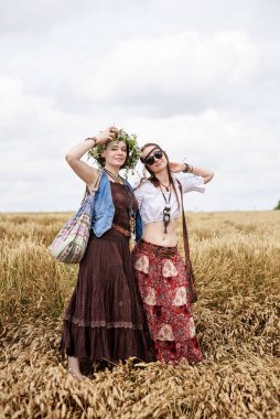İki hippi kadın, boho tarzı kıyafetler giyiyorlar, buğday tarlasında dans ediyorlar, eğleniyorlar. Kırsalda birlikte seyahat eden bayan arkadaşlar. Eko turizm konsepti. Yaz tatili boş zaman eğlencesi
