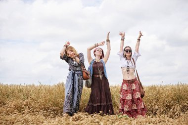 Üç hippi kadın, boho tarzı kıyafetler giyiyorlar, buğday tarlasında dans ediyorlar, eğleniyorlar. Kırsalda birlikte seyahat eden bayan arkadaşlar. Eko turizm konsepti. Yaz tatili boş zaman eğlencesi