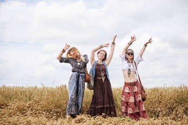 Üç hippi kadın, boho tarzı kıyafetler giyiyorlar, buğday tarlasında dans ediyorlar, eğleniyorlar. Kırsalda birlikte seyahat eden bayan arkadaşlar. Eko turizm konsepti. Yaz tatili boş zaman eğlencesi