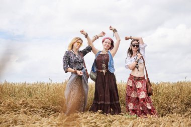 Üç hippi kadın, boho tarzı kıyafetler giyiyorlar, buğday tarlasında dans ediyorlar, eğleniyorlar. Kırsalda birlikte seyahat eden bayan arkadaşlar. Eko turizm konsepti. Yaz tatili boş zaman eğlencesi