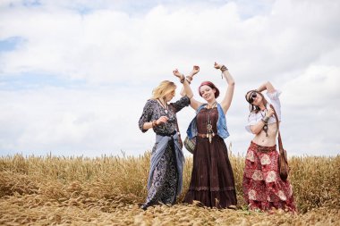 Üç hippi kadın, boho tarzı kıyafetler giyiyorlar, buğday tarlasında dans ediyorlar, eğleniyorlar. Kırsalda birlikte seyahat eden bayan arkadaşlar. Eko turizm konsepti. Yaz tatili boş zaman eğlencesi