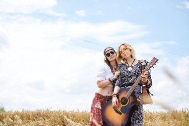 Boho tarzı kıyafetler giyen, gitar çalan ve sarı buğday tarlasında gülümseyen iki genç sıska hippi kadın. Kadın müzisyen, kırsalda müzik aleti ile poz veriyor. Yaz tatili boş zaman.