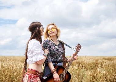 Boho tarzı kıyafetler giyen, gitar çalan ve sarı buğday tarlasında gülümseyen iki genç sıska hippi kadın. Kadın müzisyen, kırsalda müzik aleti ile poz veriyor. Yaz tatili boş zaman.