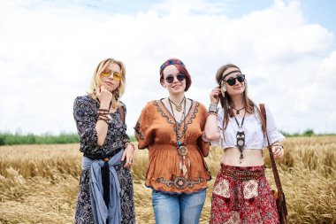 Üç hippi kadın, boho tarzı kıyafetler giyiyorlar, buğday tarlasında duruyorlar, gülümsüyorlar, kırsalda birlikte seyahat ediyorlar. Eko turizm konsepti. Yaz tatili zamanı