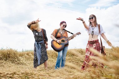 Üç hippi kadın, boho tarzı kıyafetler giyiyorlar, buğday tarlasında dans ediyorlar, gitar çalıyorlar, gülüyorlar, bayan arkadaşlar, kırsalda birlikte seyahat ediyorlar. Eko turizm konsepti. Yaz tatili zamanı