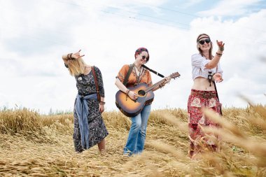 Üç hippi kadın, boho tarzı kıyafetler giyiyorlar, buğday tarlasında dans ediyorlar, gitar çalıyorlar, gülüyorlar, bayan arkadaşlar, kırsalda birlikte seyahat ediyorlar. Eko turizm konsepti. Yaz tatili zamanı