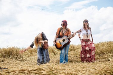 Üç hippi kadın, boho tarzı kıyafetler giyiyorlar, buğday tarlasında dans ediyorlar, gitar çalıyorlar, gülüyorlar, bayan arkadaşlar, kırsalda birlikte seyahat ediyorlar. Eko turizm konsepti. Yaz tatili zamanı
