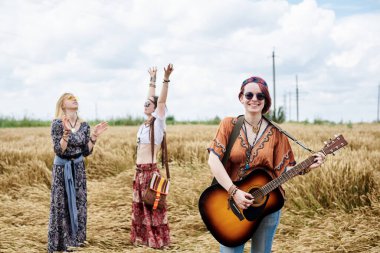 Üç hippi kadın, boho tarzı kıyafetler giyiyorlar, buğday tarlasında dans ediyorlar, gitar çalıyorlar, gülüyorlar, bayan arkadaşlar, kırsalda birlikte seyahat ediyorlar. Eko turizm konsepti. Yaz tatili zamanı