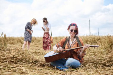 Kızıl saçlı hippi kadınlar, boho tarzı kıyafetler giyiyorlar, buğday tarlasında oturuyorlar, arka planda iki kız arkadaşıyla gitar çalıyorlar. Kırsalda birlikte seyahat etmek. Çevre turizmi. Yaz tatili