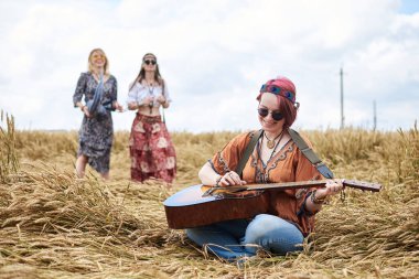 Kızıl saçlı hippi kadınlar, boho tarzı kıyafetler giyiyorlar, buğday tarlasında oturuyorlar, arka planda iki kız arkadaşıyla gitar çalıyorlar. Kırsalda birlikte seyahat etmek. Çevre turizmi. Yaz tatili