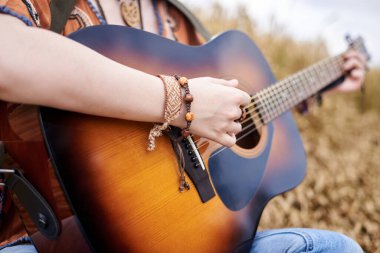 Kızıl saçlı, boho tarzı kıyafetler giyen, buğday tarlasının ortasında oturan, kırsalda gezen müzisyen genç hippi kadınların elinde tutulan akustik gitarın yakın plan fotoğrafı..