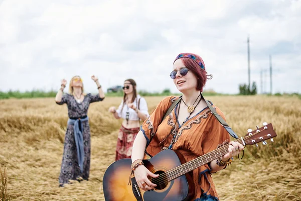 Three hippie women, wearing boho style clothes, dancing in the wheat field, playing guitar, laughing, Female friends, traveling together in countryside. Eco tourism concept. Summer leisure time