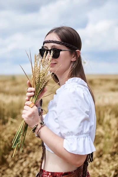 年轻女子 棕色长发 身穿嬉皮衣 站在麦田中央 拿着小麦茎束 自然背景的富有创意的女性肖像 生态旅游概念 — 图库照片