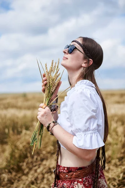 年轻女子 棕色长发 身穿嬉皮衣 站在麦田中央 拿着小麦茎束 自然背景的富有创意的女性肖像 生态旅游概念 — 图库照片