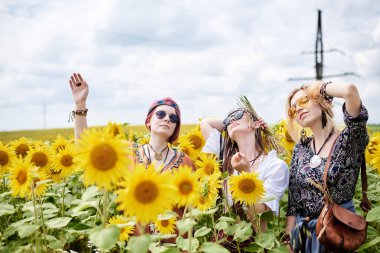 Üç genç kadın, boho hippi kıyafetleri giyiyorlar, sarı ayçiçeği tarlasının ortasında duruyorlar. Kız arkadaşlar, kırsalda birlikte seyahat ediyorlar. Çevre turizmi kavramı.