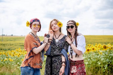 Üç genç kadın, boho hippi kıyafetleri giyiyorlar, sarı ayçiçeği tarlasının ortasında duruyorlar. Kız arkadaşlar, kırsalda birlikte seyahat ediyorlar. Çevre turizmi kavramı.