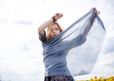Genç sarışın kadın, boho hippi kıyafetleri giyiyor, gri şeffaf şal eşarbı tutuyor, sarı ayçiçeği tarlasında rüzgarda sallanıyor. Doğal arka planda yaratıcı kadın portresi. Çevre turizmi kavramı.