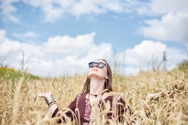 Genç esmer hippi kadın, boho tarzı kıyafetler giyiyor, mavi gökyüzüyle buğday tarlasının ortasında oturuyor, meditasyon yapıyor, düşünüyor, rahatlıyor, eğleniyor. Eko turizm konsepti. Doğa koruması.