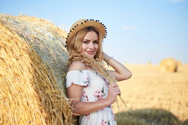 Genç sarışın kadın, uzun beyaz romantik elbise ve hasır şapka giyiyor, yazın tarlada buğday balyasına yaslanıyor. Doğal arka planda kadın portresi.