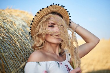 Genç sarışın kadın, uzun beyaz romantik elbise ve hasır şapka giyiyor, yazın tarlada buğday balyasına yaslanıyor. Doğal arka planda kadın portresi.