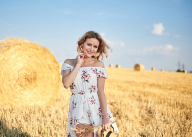 Sarışın, uzun, romantik bir elbise giyen, elinde hasır şapka ve kurutulmuş çimen buketi tutan, yazın tarlada buğday balyasının önünde poz veren genç bir kadın. Doğal arka planda kadın portresi.