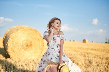 Sarışın, uzun, romantik bir elbise giyen, elinde hasır şapka ve kurutulmuş çimen buketi tutan, yazın tarlada buğday balyasının önünde poz veren genç bir kadın. Doğal arka planda kadın portresi.