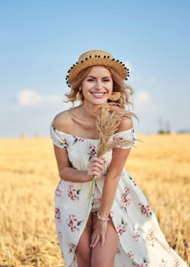 Sarışın, uzun, romantik bir elbise ve hasır şapka giyen, elinde kurutulmuş çim buketi tutan yazın tarlada buğday balyasının önünde poz veren genç bir kadın. Doğal arka planda kadın portresi.