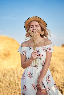 Sarışın, uzun, romantik bir elbise ve hasır şapka giyen, elinde kurutulmuş çim buketi tutan yazın tarlada buğday balyasının önünde poz veren genç bir kadın. Doğal arka planda kadın portresi.