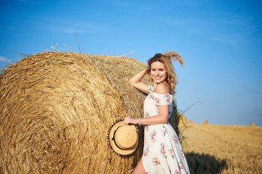 Genç sarışın kadın, uzun beyaz romantik elbise ve hasır şapka giyiyor, yazın tarlada buğday balyasına yaslanıyor. Doğal arka planda kadın portresi.
